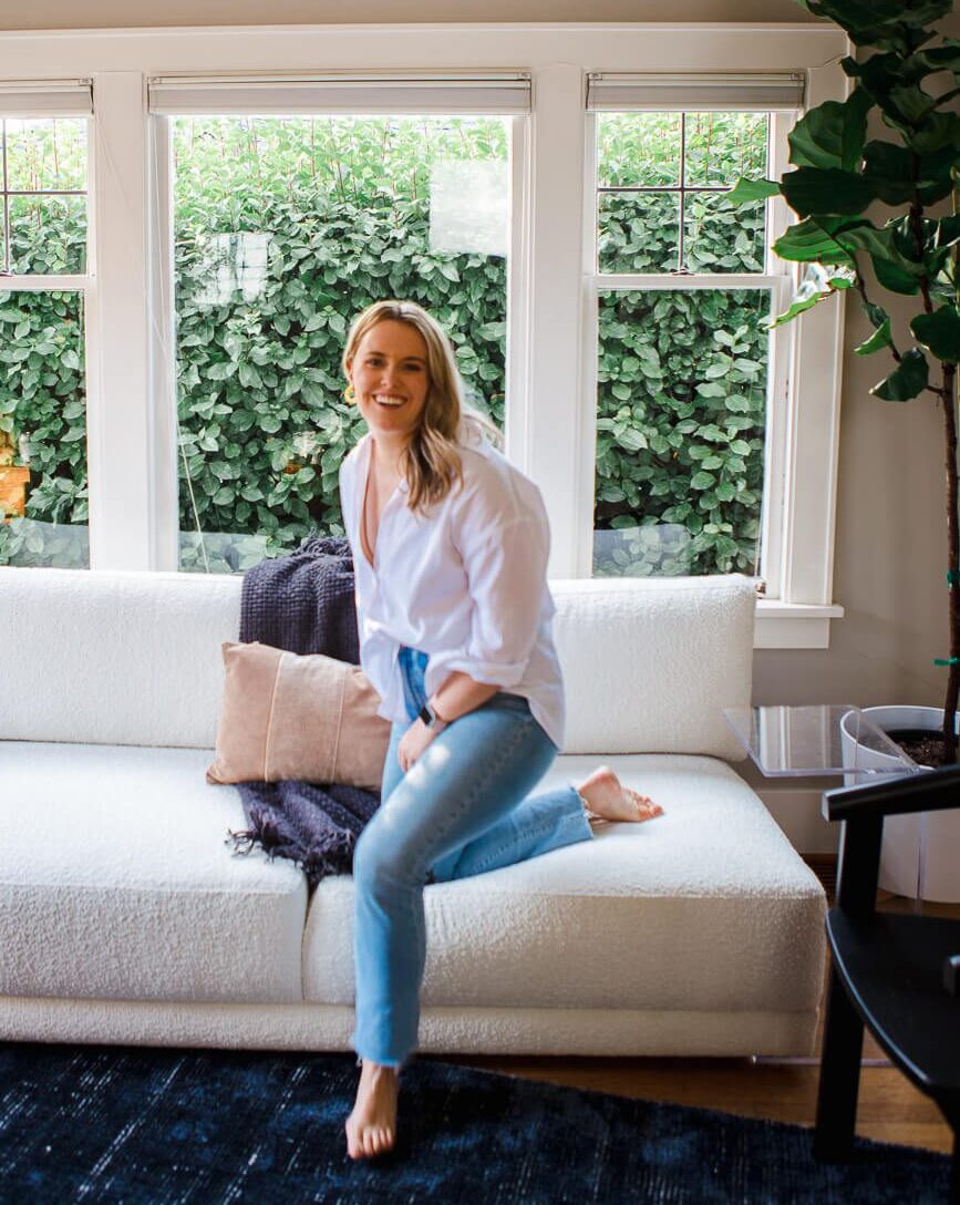 Photo of Alex Alexander, Half standing with one knee on the couch. She has blonde hair and is looking at the camera laughing. Wearing a white longsleeve button down shirt and blue jeans.