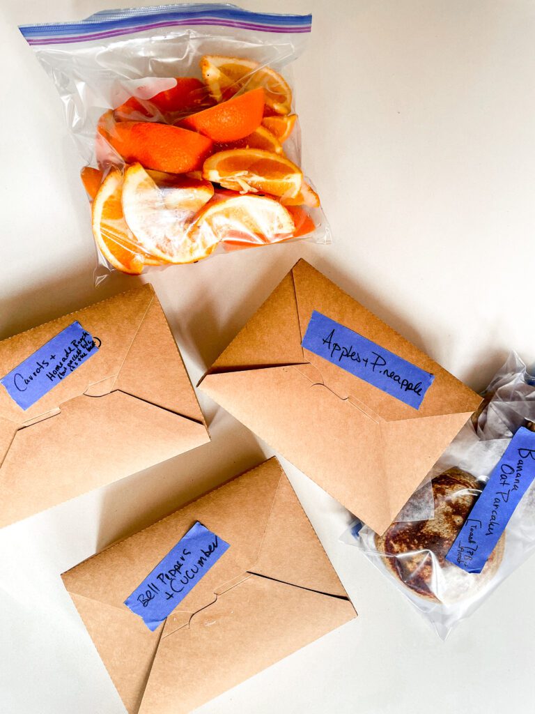 Brown to-go boxes packed with foods and labelled - apples and pineapple, carrots, bell peppers and cucumbers. A ziplock full of pancakes and a bag of sliced oranges. 