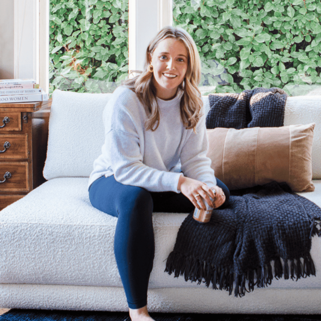 Alex Alexander, a blonde hair female wearing leggings and a white sweater, is sitting on a large white sofa in front of big windows.