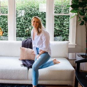 Photo of Alex Alexander, Half standing with one knee on the couch. She has blonde hair and is looking at the camera laughing. Wearing a white longsleeve button down shirt and blue jeans.